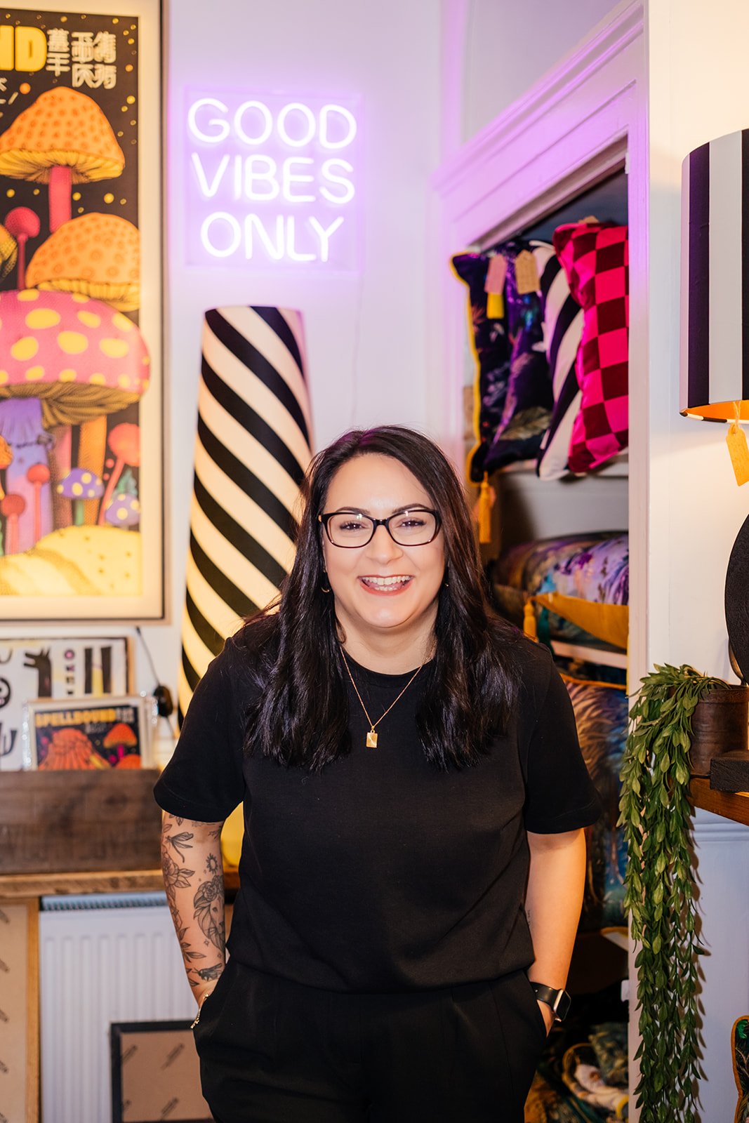 A woman with glasses and long dark hair smiling in a colorful boutique or shop with vibrant mushrooms art, a neon sign saying 'Good Vibes Only,' and shelves of pillows and fabrics.