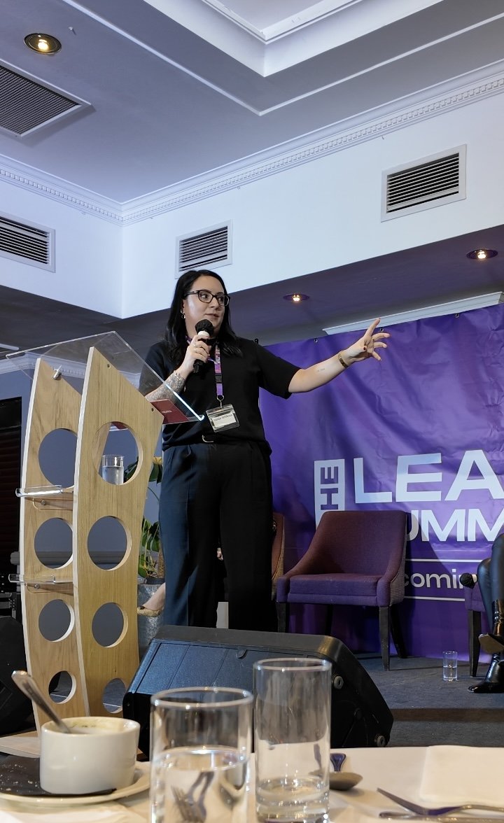A woman speaking on stage at a conference or seminar, holding a microphone and gesturing with her hand, with a purple banner in the background that says 'The Lead Summit'.